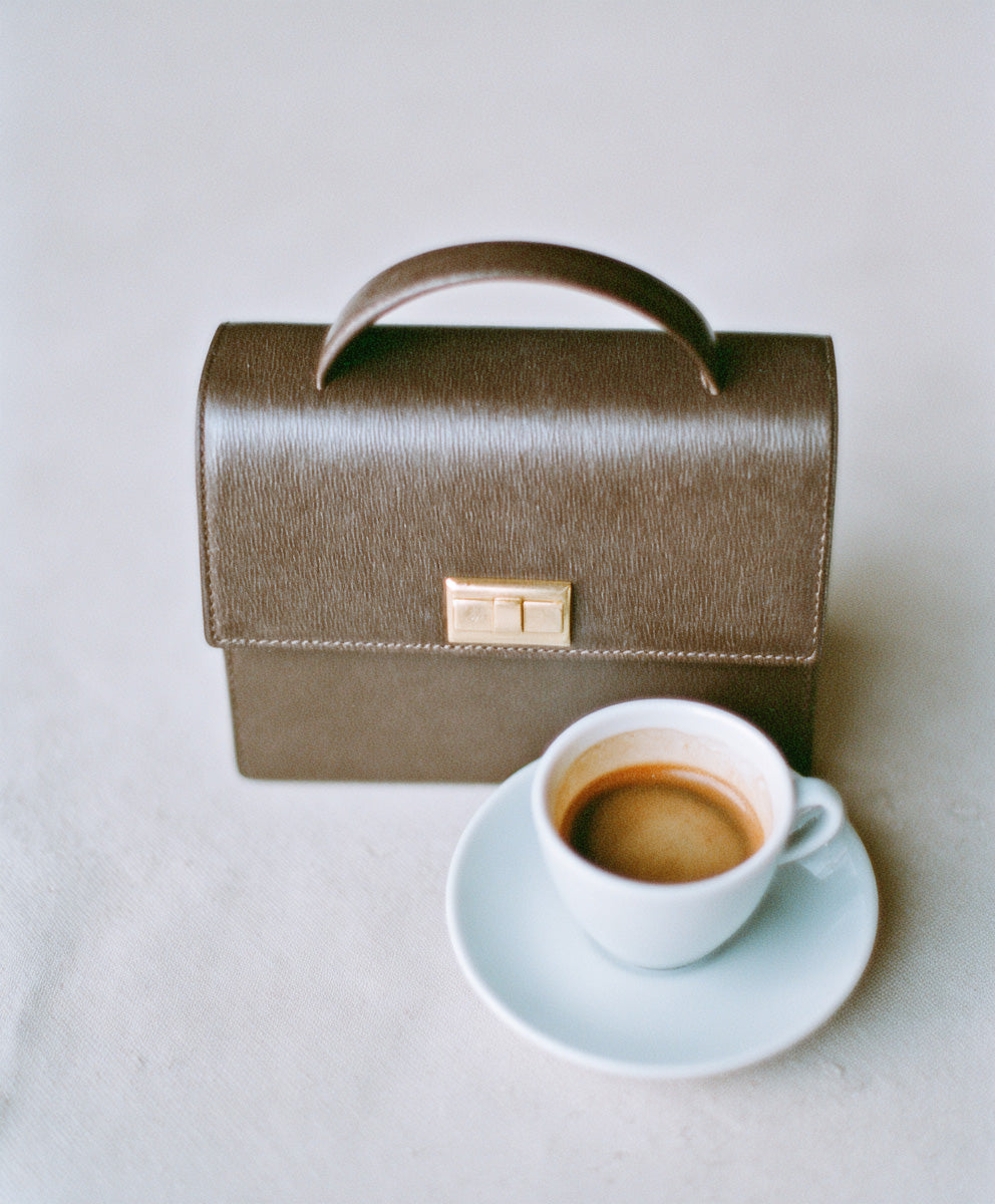 Close-up of a structured brown leather handbag with gold clasp placed beside a white cup of espresso on a saucer.