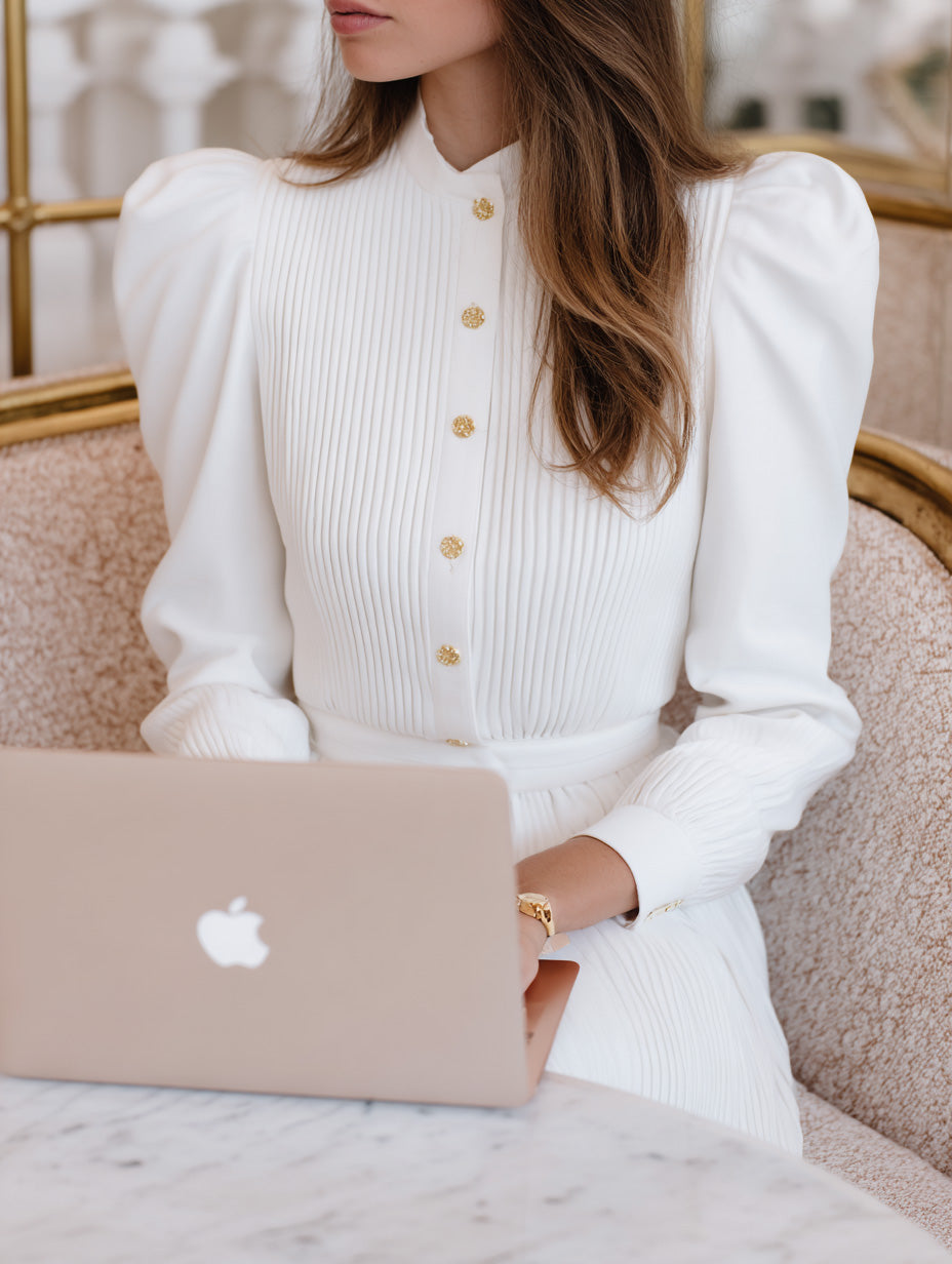 Woman in elegant white pleated dress working on laptop at marble table, editorial business stock photo