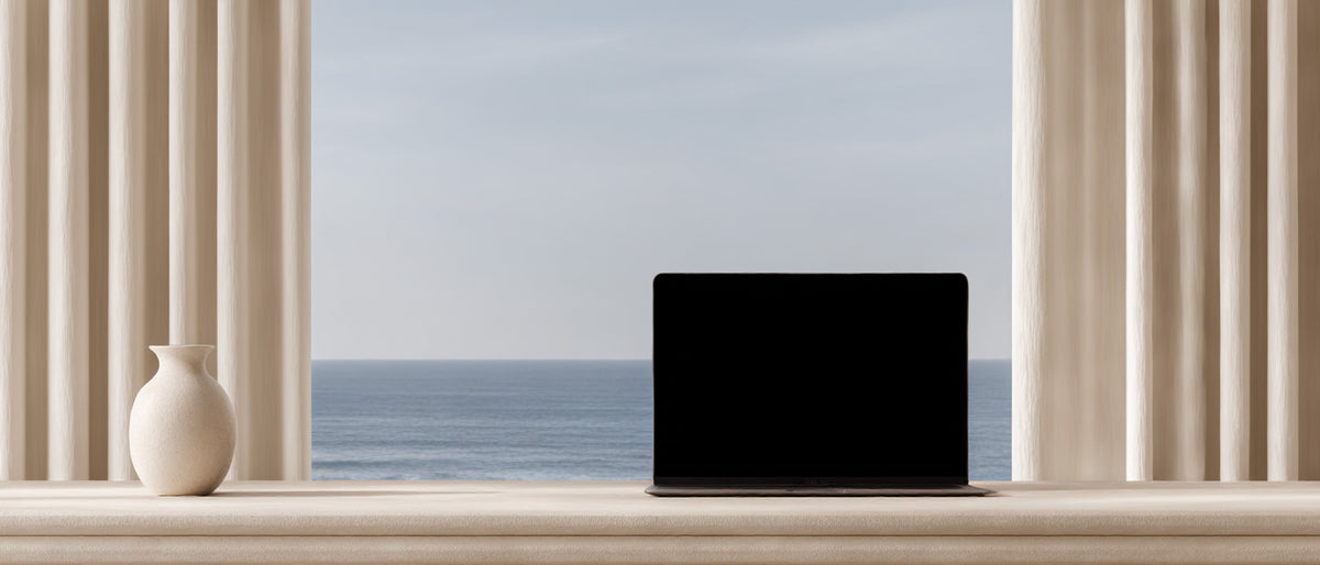 Editorial still life of a minimalist workspace with a laptop and ceramic vase placed before a window with an ocean view.