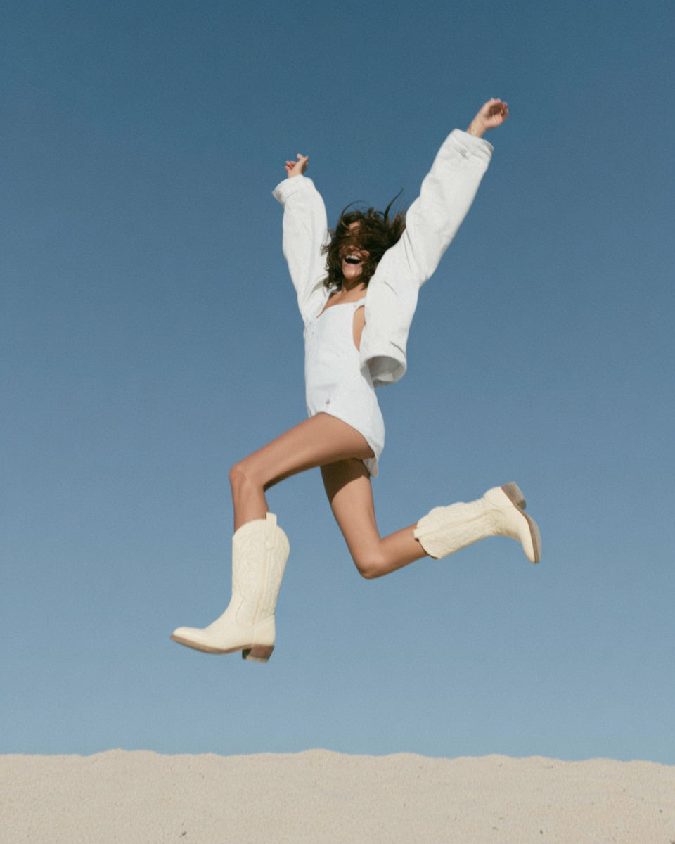 Editorial photo of a woman in a white outfit and boots jumping joyfully against a clear blue sky.